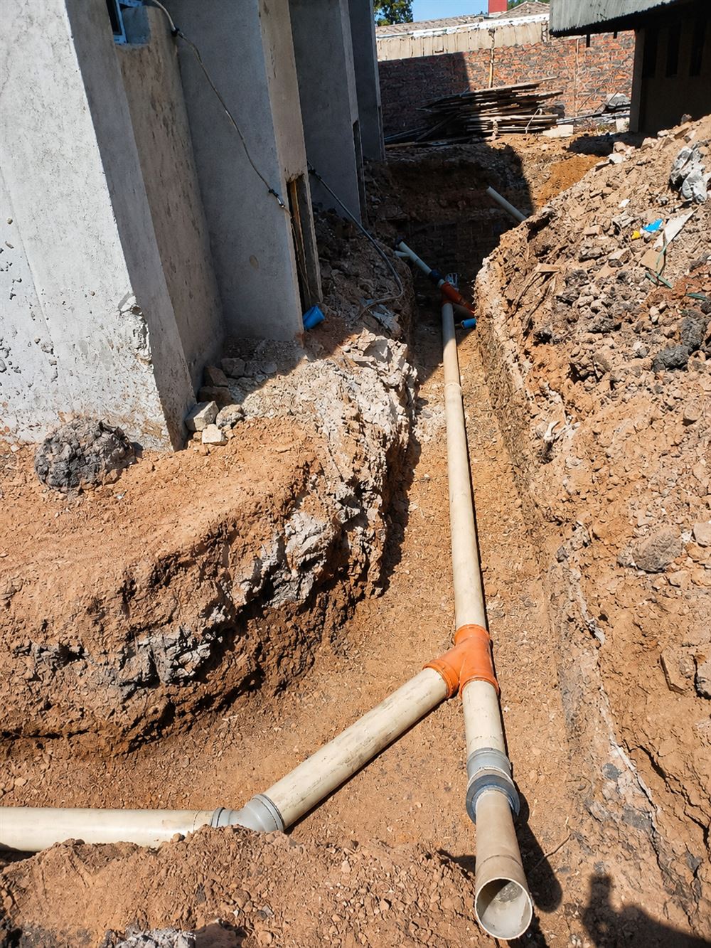 PVC soil pipes meeting at a junction fitting in an open trench beside a building under construction